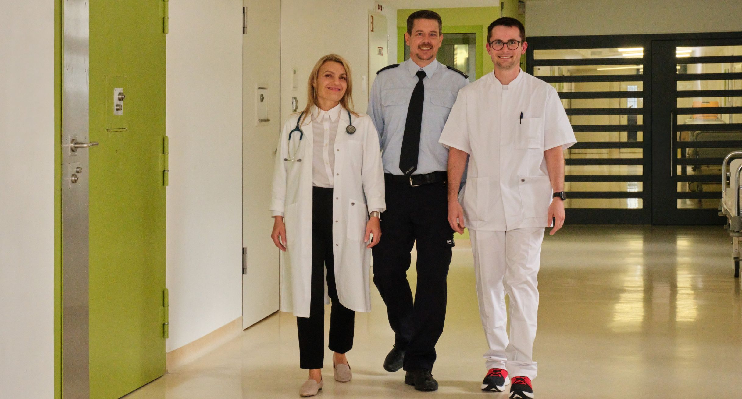 Ärztin, Pfleger und ein Vollzugsbediensteter laufen durch eine Krankenstation.