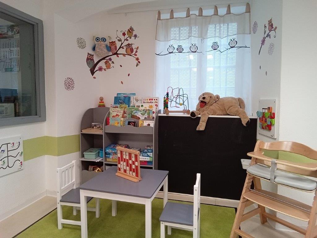 Spielecke im Besuch. Weiße Wand mit grünem Streifen und Eulenbild an der Wand. Gründer Teppich und ein kleiner Tisch mit zwei Stühlen und einem Hochstuhl. In einem Regal Bücher und Spielsachen.