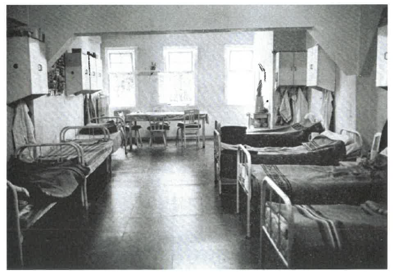 Schwarz-Wei&szlig;-Bild einer Krankenzelle mit sieben Betten und einem Tisch am Fenster.